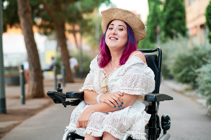 Young woman who uses a wheelchair smiles at camera on a city pavement.