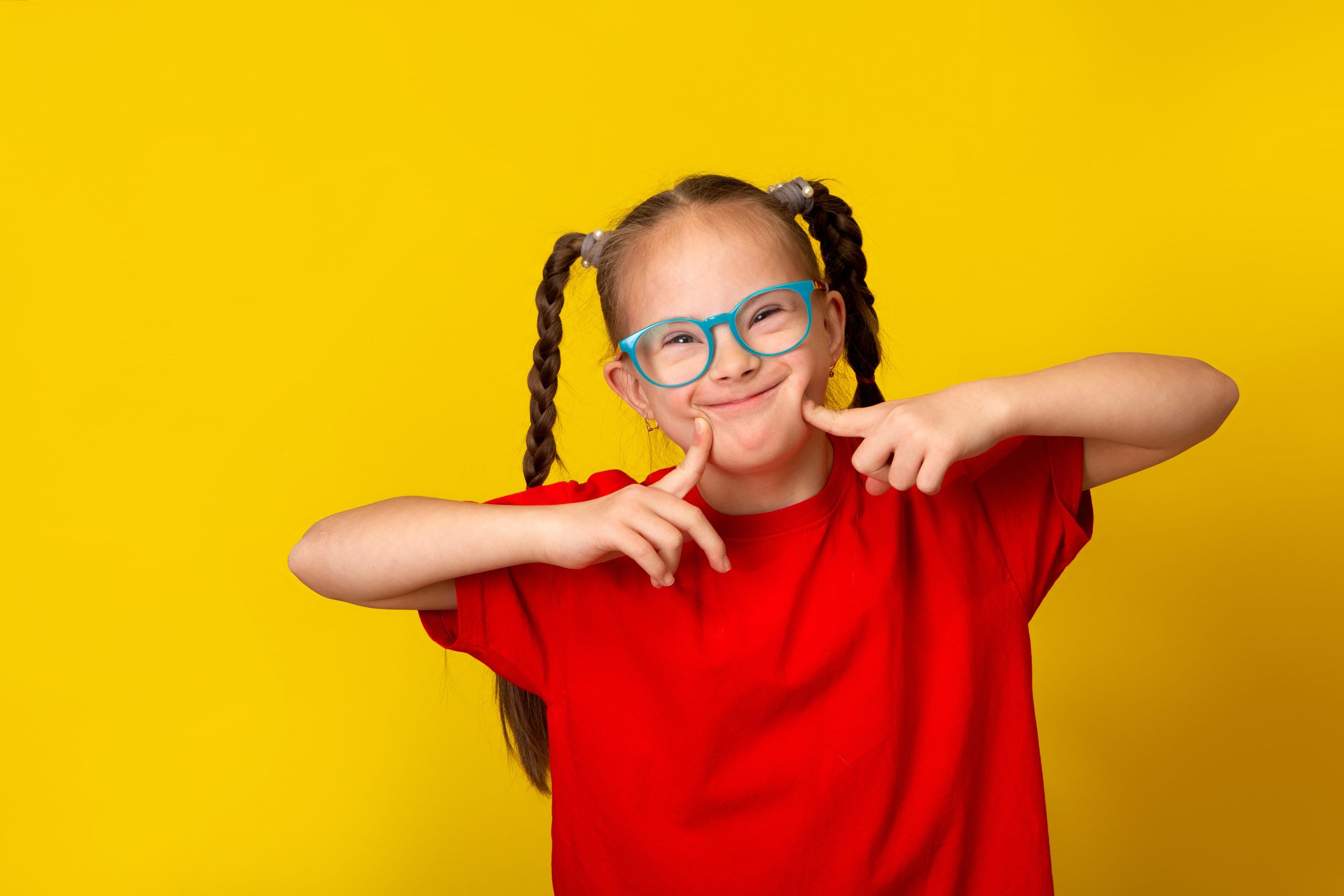 Young girl with Downs Syndrome smiles at the camera against a bright yellow background.